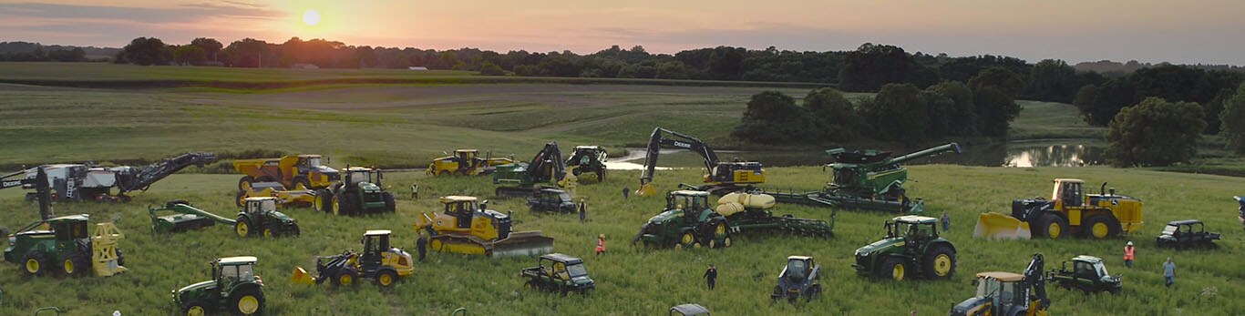 Хора, които се разхождат сред няколко вида оборудване на John Deere в голямо поле.