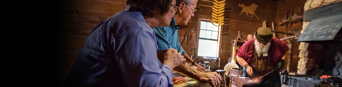 Възрастна двойка, която гледа ковач как кове парче метален чук на историческия обект на Гранд Детауър, Илинойс, САЩ.