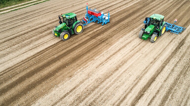 Два трактора John Deere 6M на голо поле. Единият тегли сеялка Monsomen. Другият тегли комбинирана почвообработваща машина Lemken