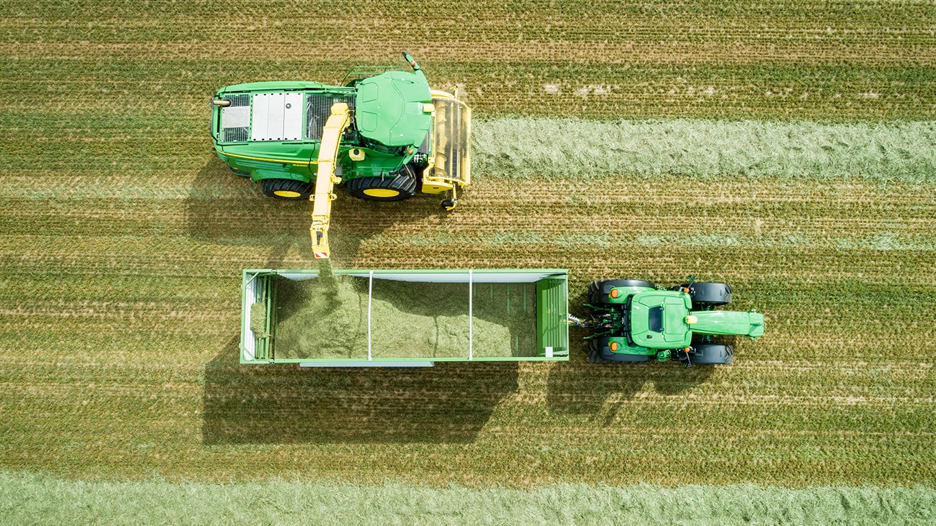 Снимка от въздуха на самоходен силажокомбайн серия 8000 на John Deere с подбирач за сено, товарещ слама на платформа, теглена от трактор на John Deere