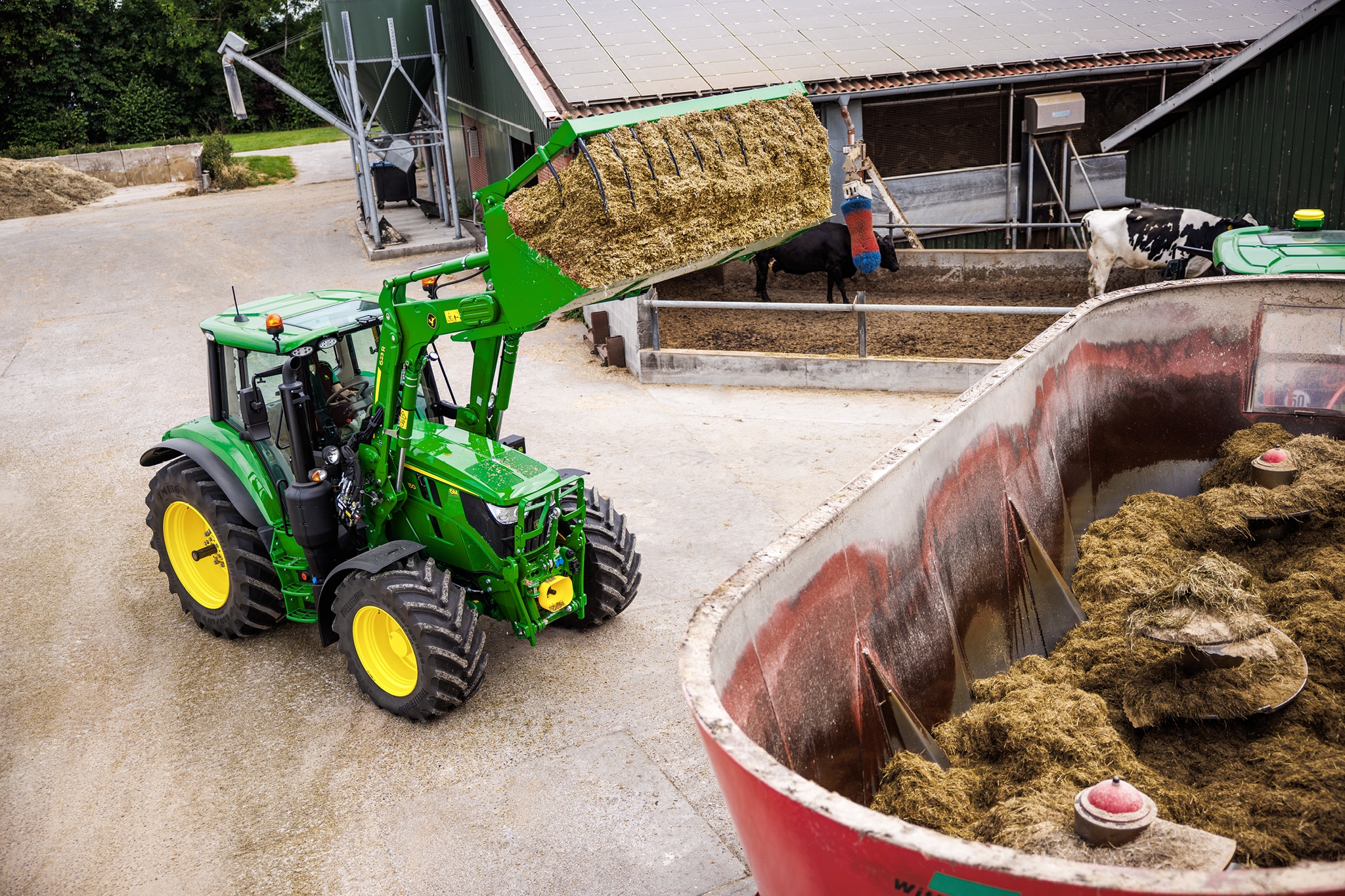 Трактор John Deere от серия 6M, използващ челен товарач за подаване на силаж в смесително прикачно приспособление в стопанство.