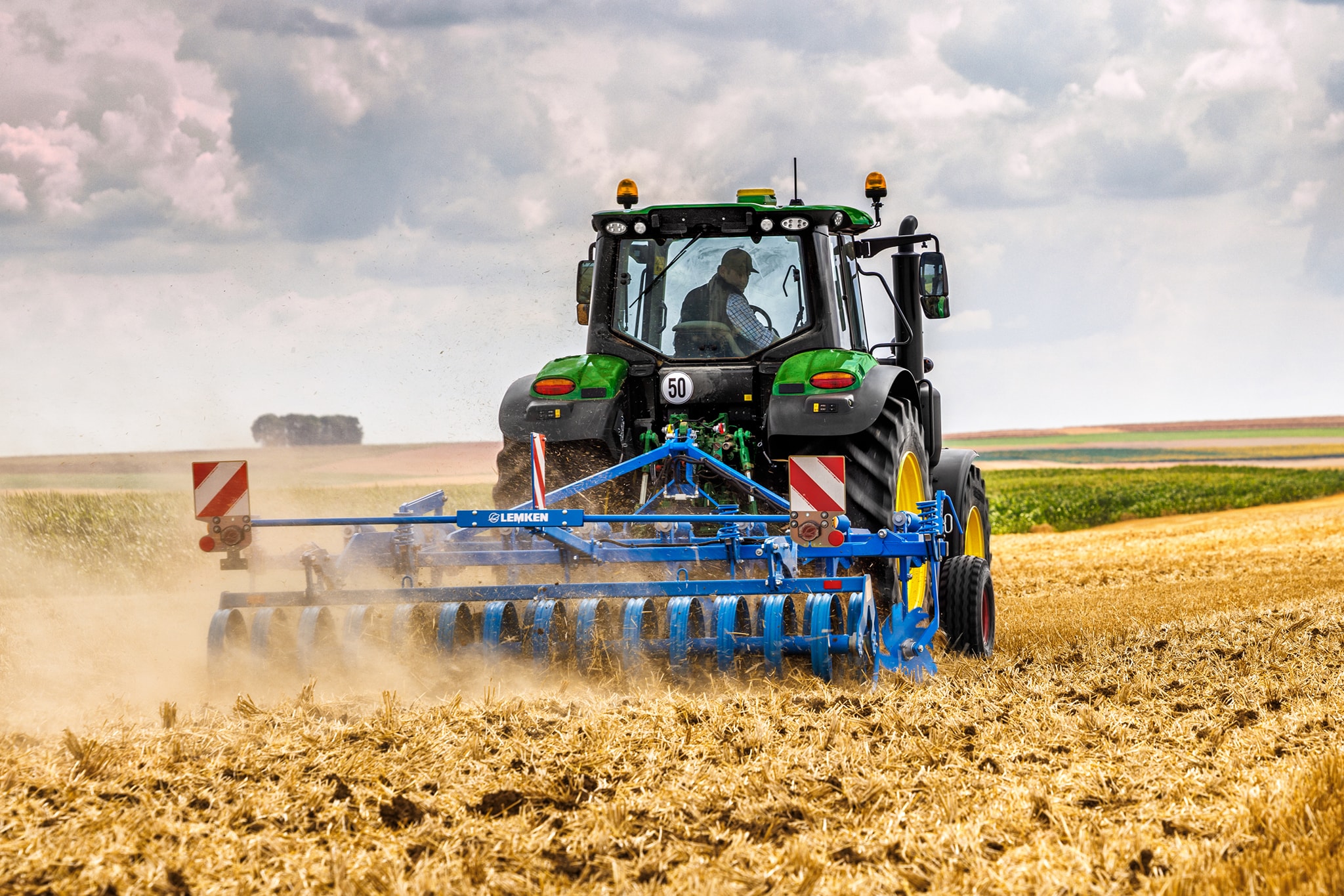Трактор John Deere от серия 6M, култивиращ ожънато поле с дискова брана, заден изглед в прашни полеви условия.