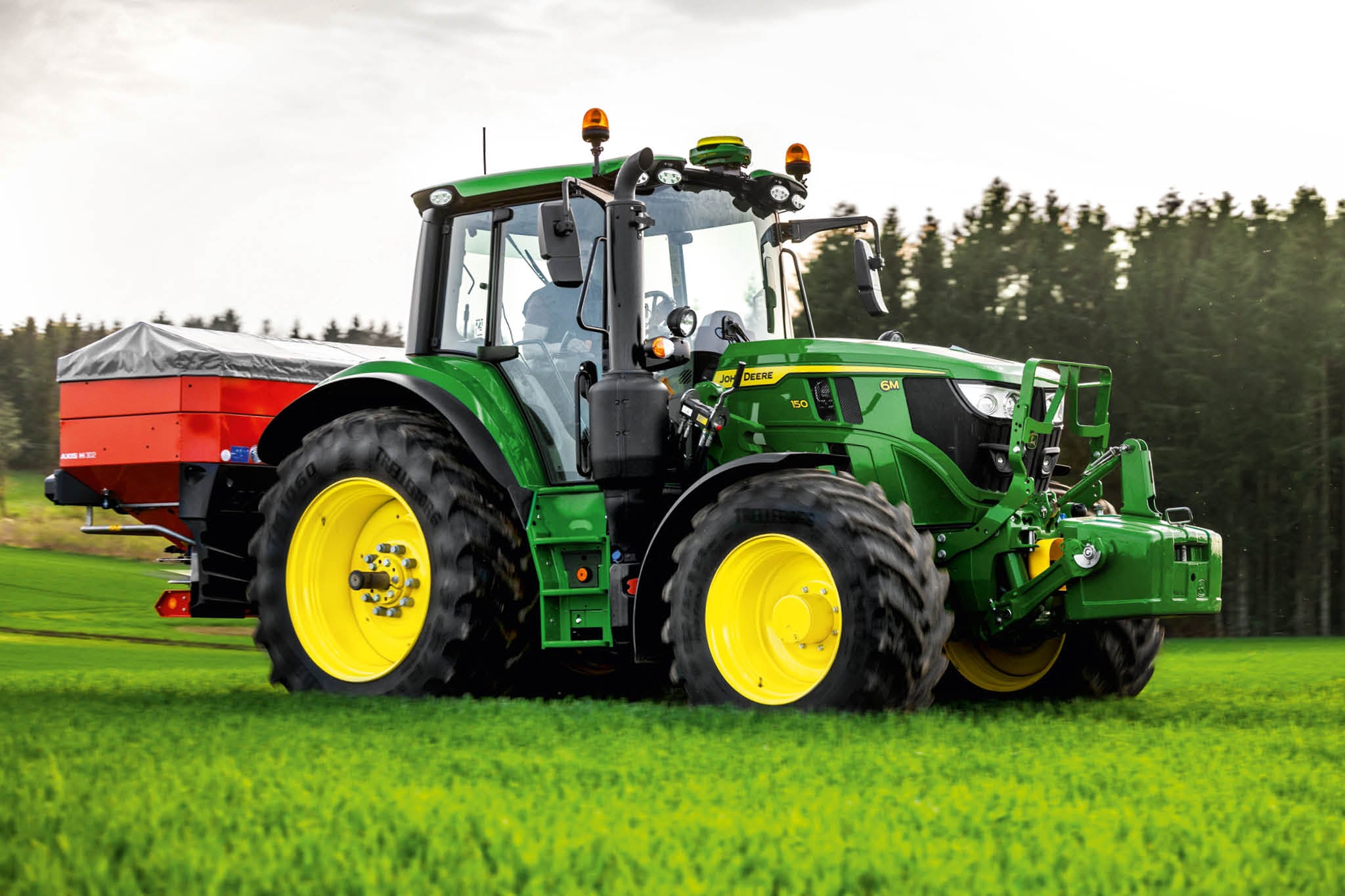 Трактор John Deere от серия 6M със заден разпръсквач, работещ върху трева близо до ръба на гората.