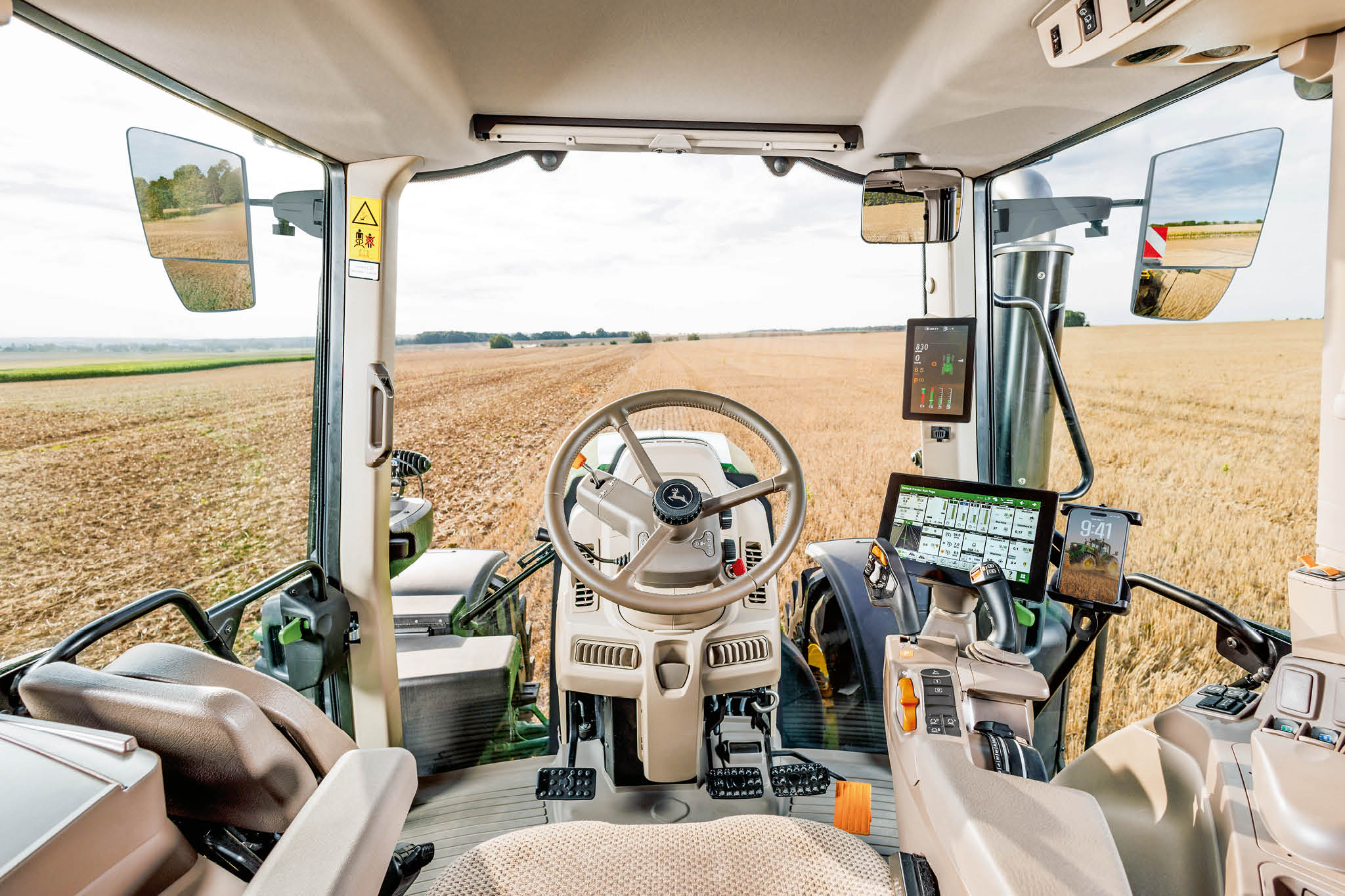 Вътрешността на кабината на трактора на John Deere с волан, дисплеи за цифрово управление и изглед на полето през предното стъкло по време на земеделска работа.