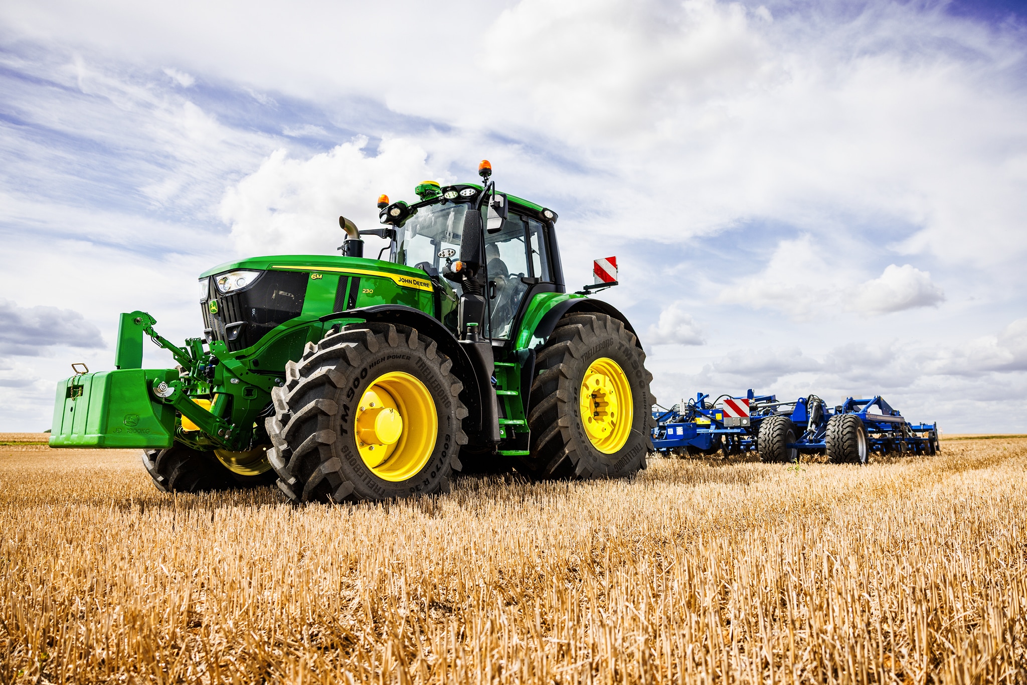 Трактор John Deere от серия 6M, който тегли тежък прикачен инвентар за оран през стърнище под облачно небе.