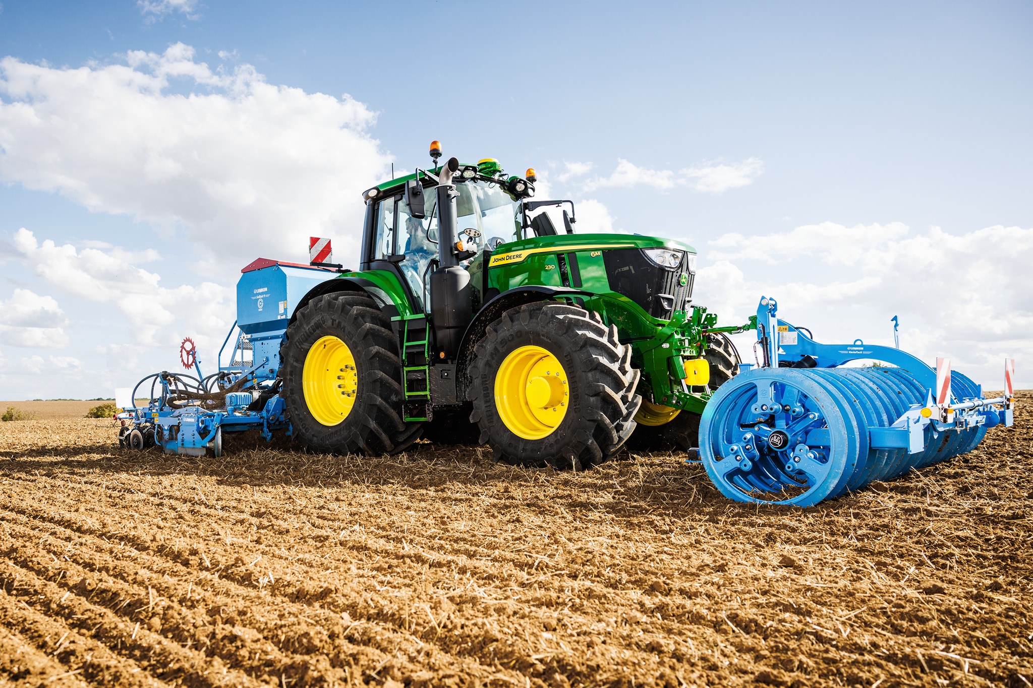 Трактор John Deere от серия 6M обработва поле с прикачен голям син ролков инвентар.