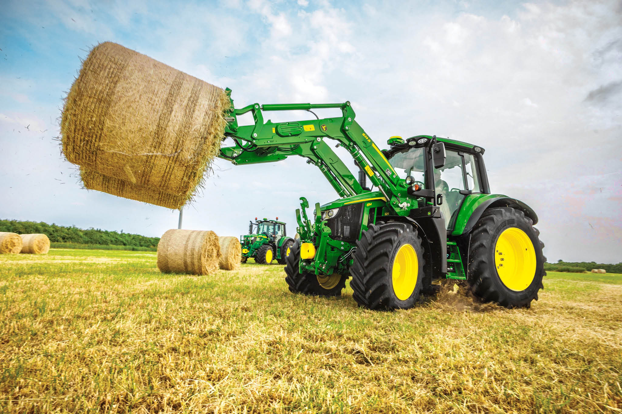Тракторът John Deere от серия 6M повдига голяма кръгла бала сено с челен товарач на открито поле.