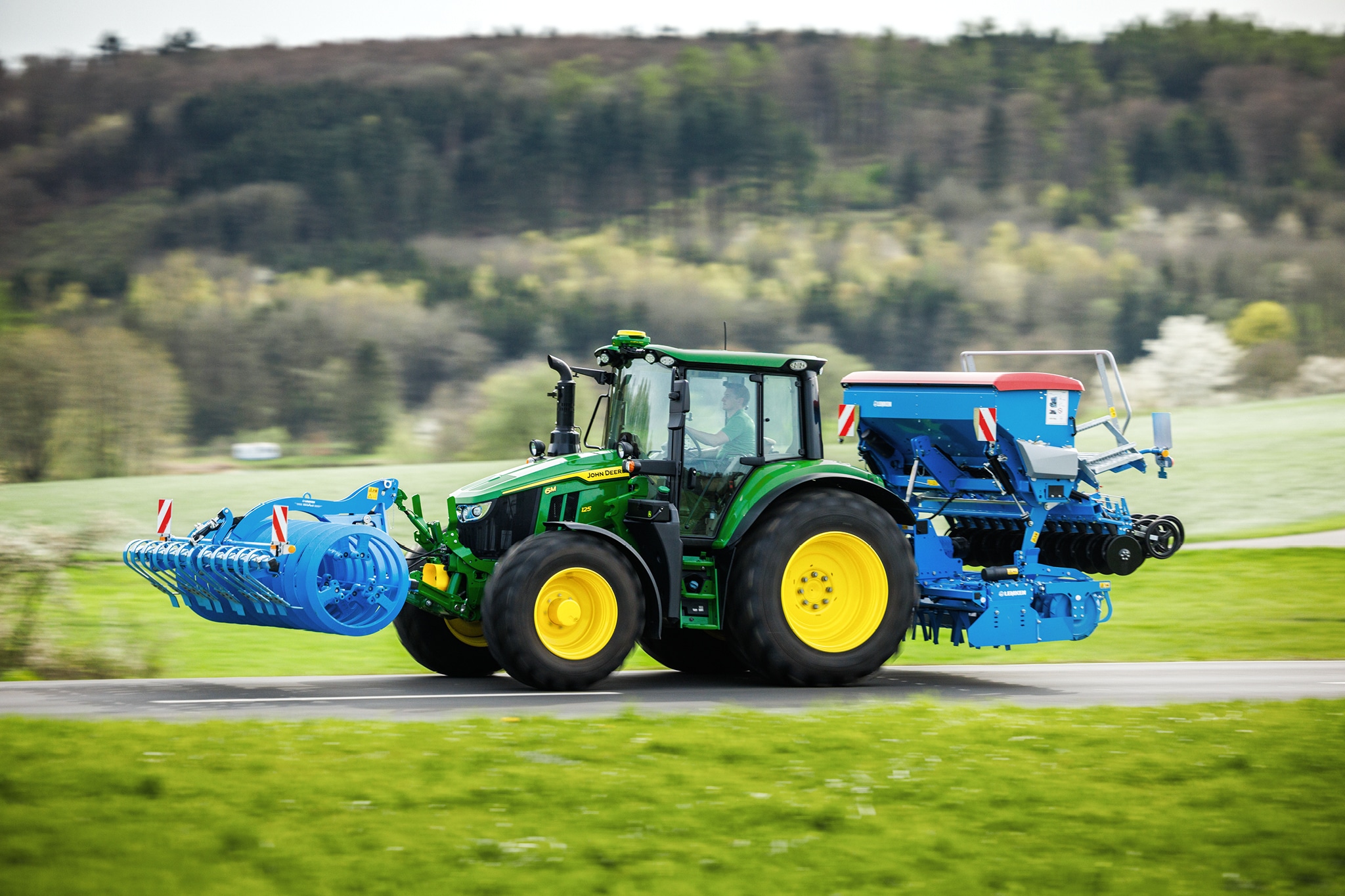 Трактор John Deere от серия 6M, който транспортира предно монтиран инвентар и задна сеялка по селски път.