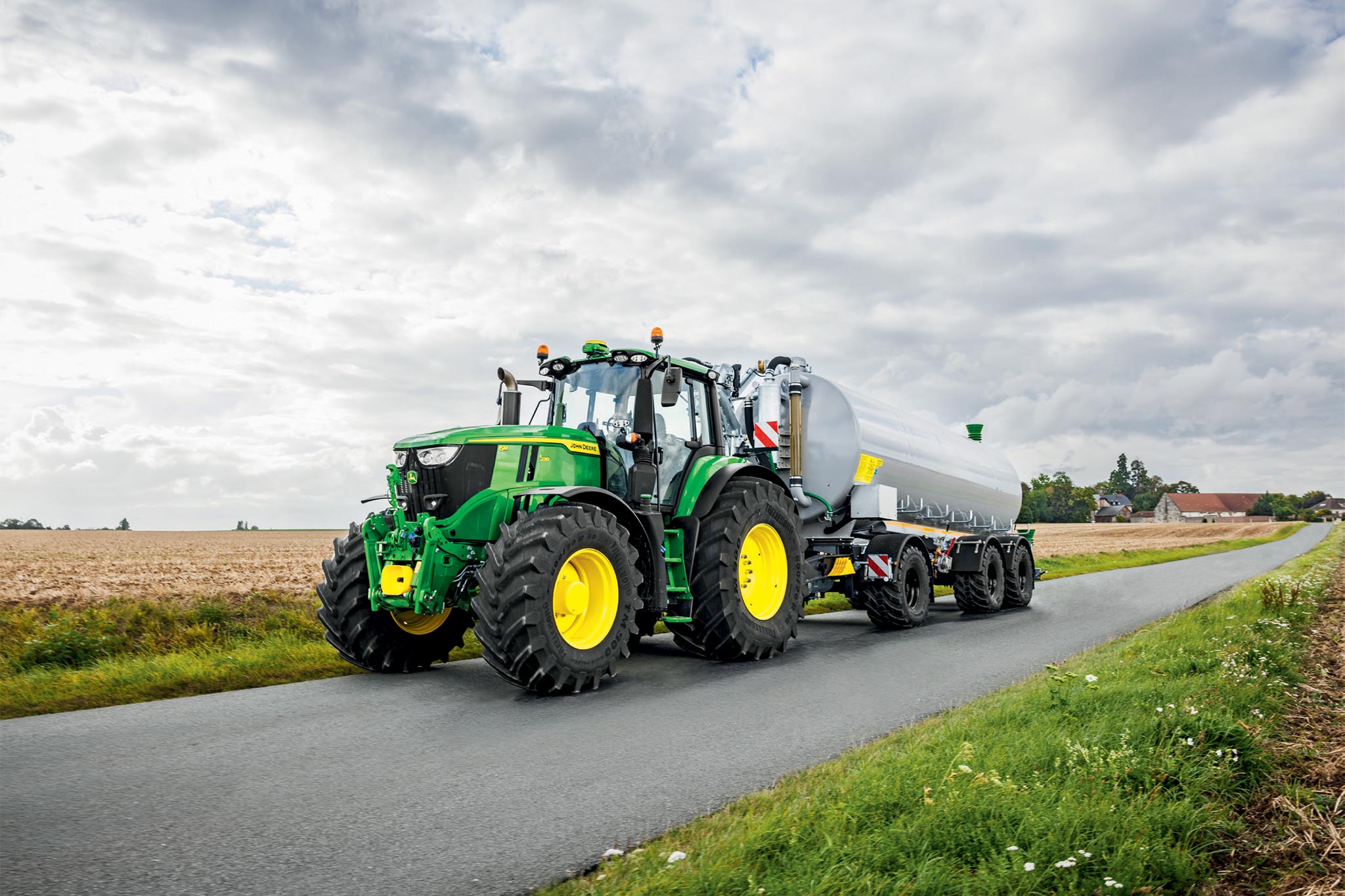Трактор John Deere от серия 6M, работещ в ожънато поле с широки култиватори, гледани отгоре, с вдигащ се прах.