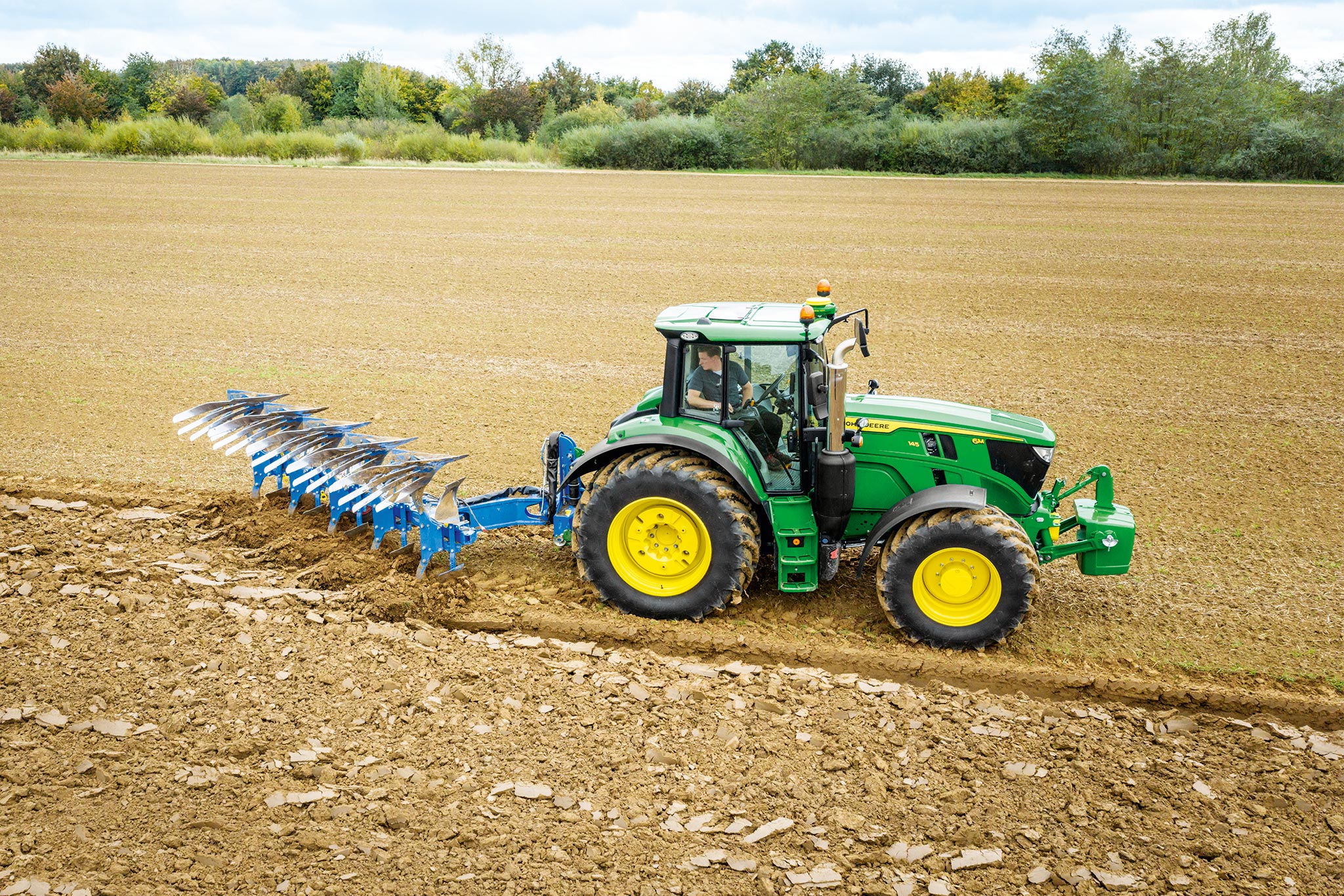 Трактор John Deere от серия 6M, работещ в разорано поле с прикачен отзад инвентар за оран под облачно небе.