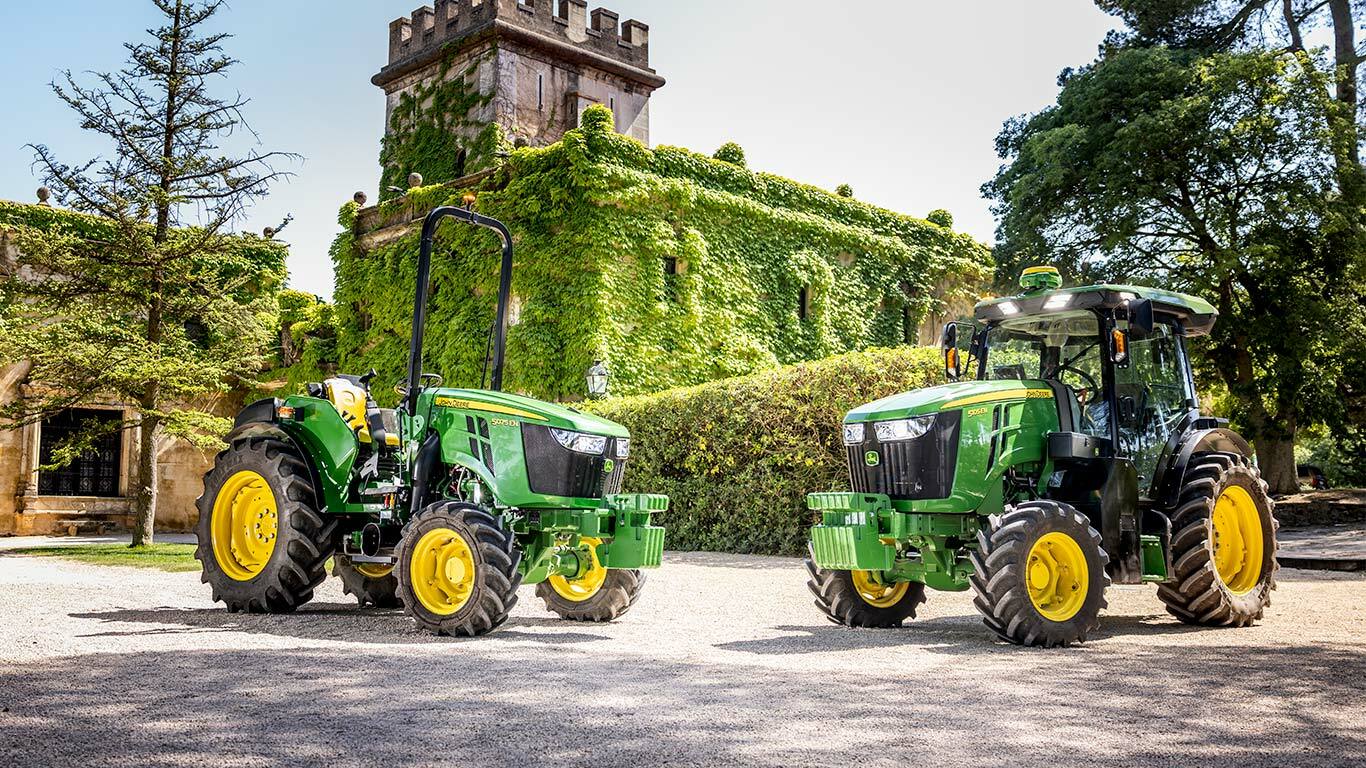 Зеленo и жълт трактор John Deere 5075EN, движещ се по черен път в овощна градина, заобиколен от плодни дръвчета и слънчева светлина.