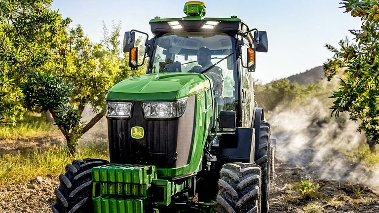 Зеленo и жълт трактор John Deere 5075EN, движещ се по черен път в овощна градина, заобиколен от плодни дръвчета и слънчева светлина.