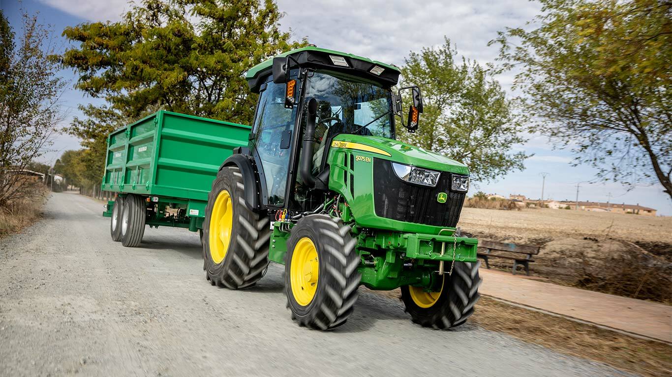 Трактор John Deere 5075EN тегли зелена ремарке по селски черен път с дървета на заден план.