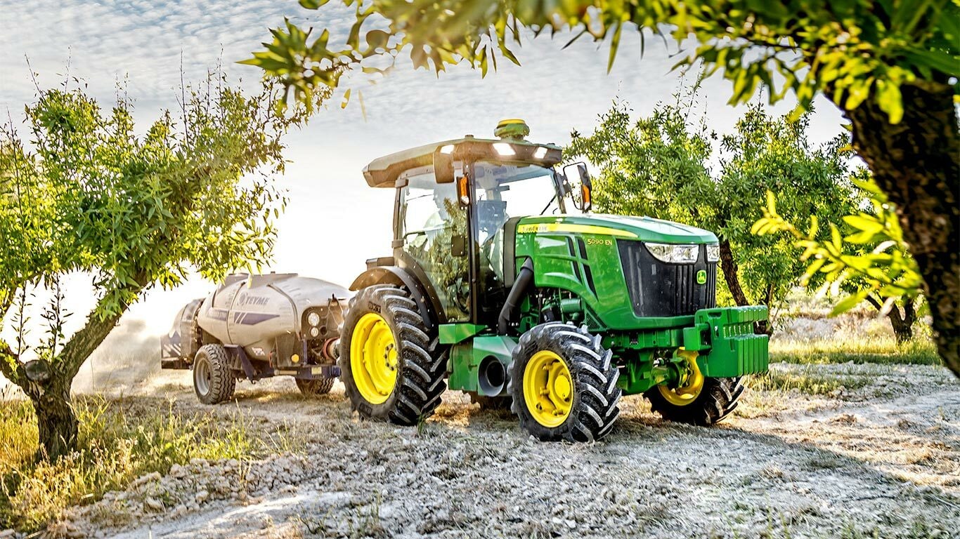 Зелен и жълт трактор John Deere 5090EN, който тегли пръскачка през овощна градина в слънчев ден.