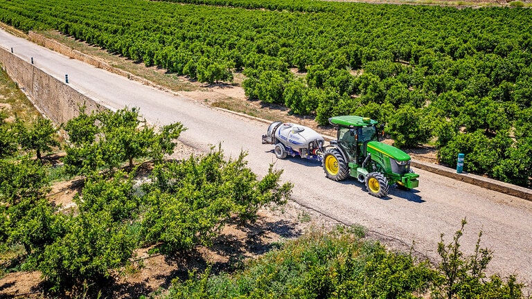 Зелен и жълт трактор John Deere 5090EN, който тегли пръскачка на павиран път между големи редове овощни градини под чисто синьо небе.