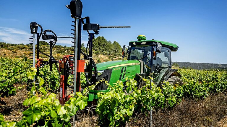 Зелен и жълт трактор John Deere 5090EN, оборудван с инструменти за подрязване, работещи в лозови масиви под чисто синьо небе.