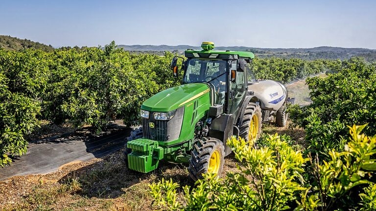 Зеленo и жълт трактор John Deere 5105EN, теглещ пръскачка в плодородна овощна градина с хълмове на заден план.