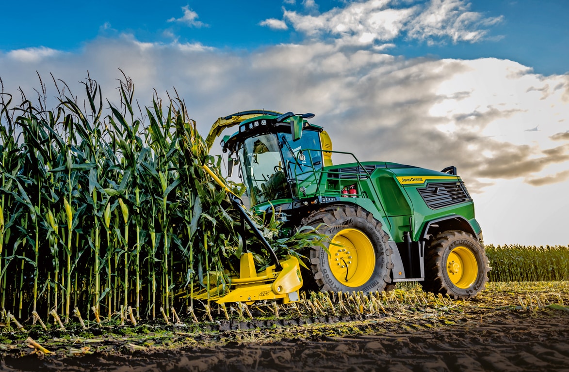 Фуражокомбайн John Deere прибира царевица в поле под синьо небе с облаци.