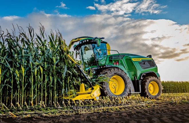 Фуражокомбайн John Deere прибира царевица в поле под синьо небе с облаци.
