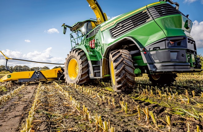 Работата на фуражокомбайн John Deere в царевично поле, където се виждат големите гуми и режещият механизъм на машината на фона на ясно синьо небе.