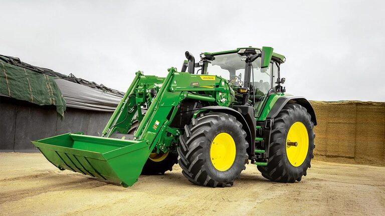 Трактор John Deere с челен товарач, паркиран върху земеделска повърхност близо до съхранение на силаж.