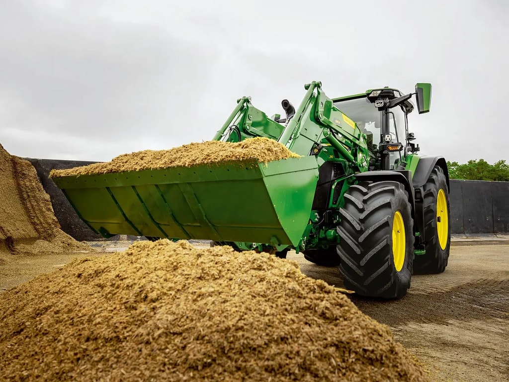 Трактор John Deere, използващ челен товарач за превоз на нарязан силаж в земеделска зона за съхранение.